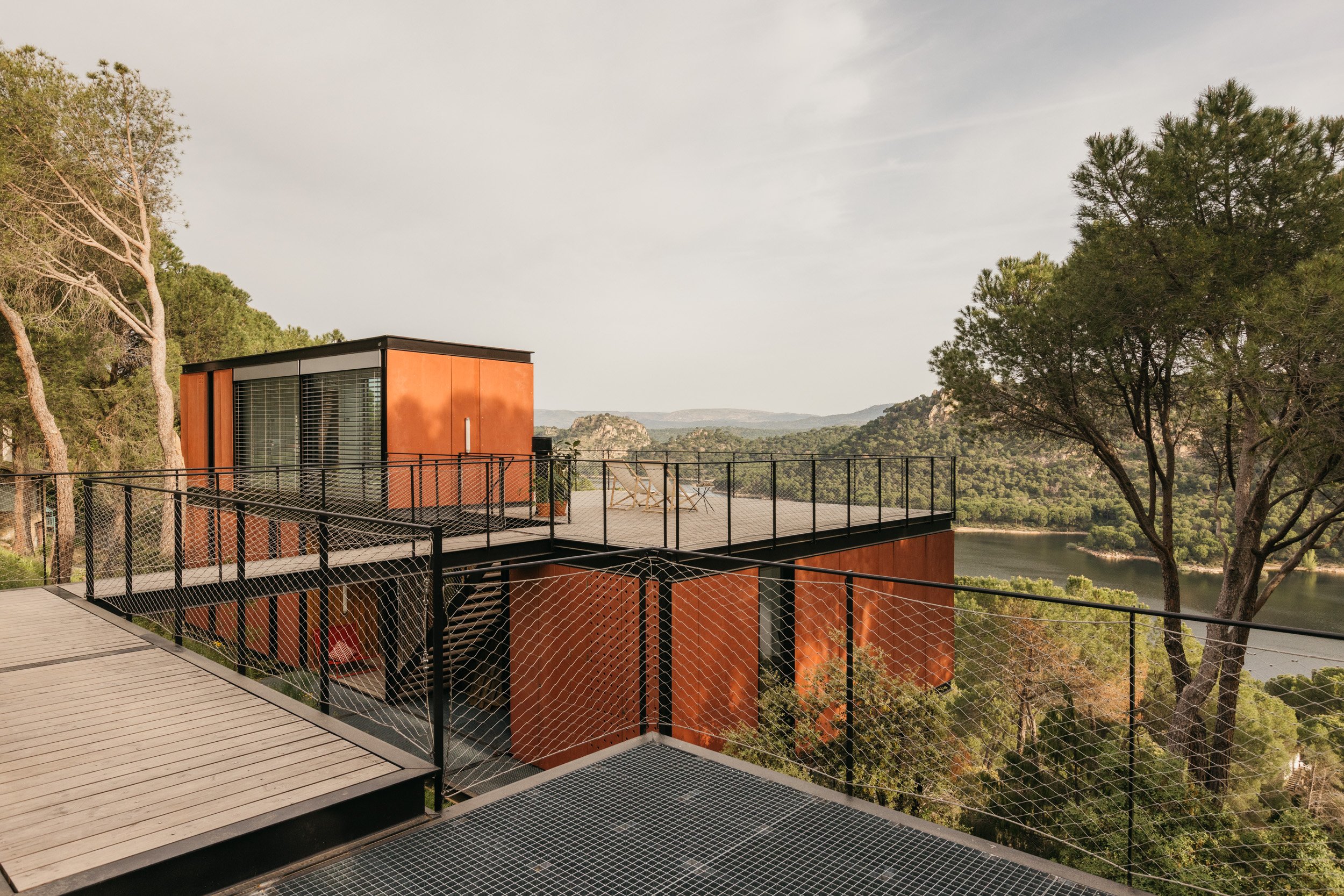 Vivienda modular Tini Case Study de acero corten en dos niveles, con terrazas y vistas a un embalse entre pinos