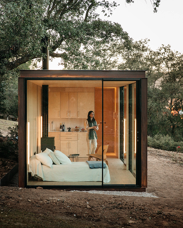 Casa modular pequeña Tini XS con fachada de cristal, dormitorio iluminado y cocina de madera en un bosque al atardecer