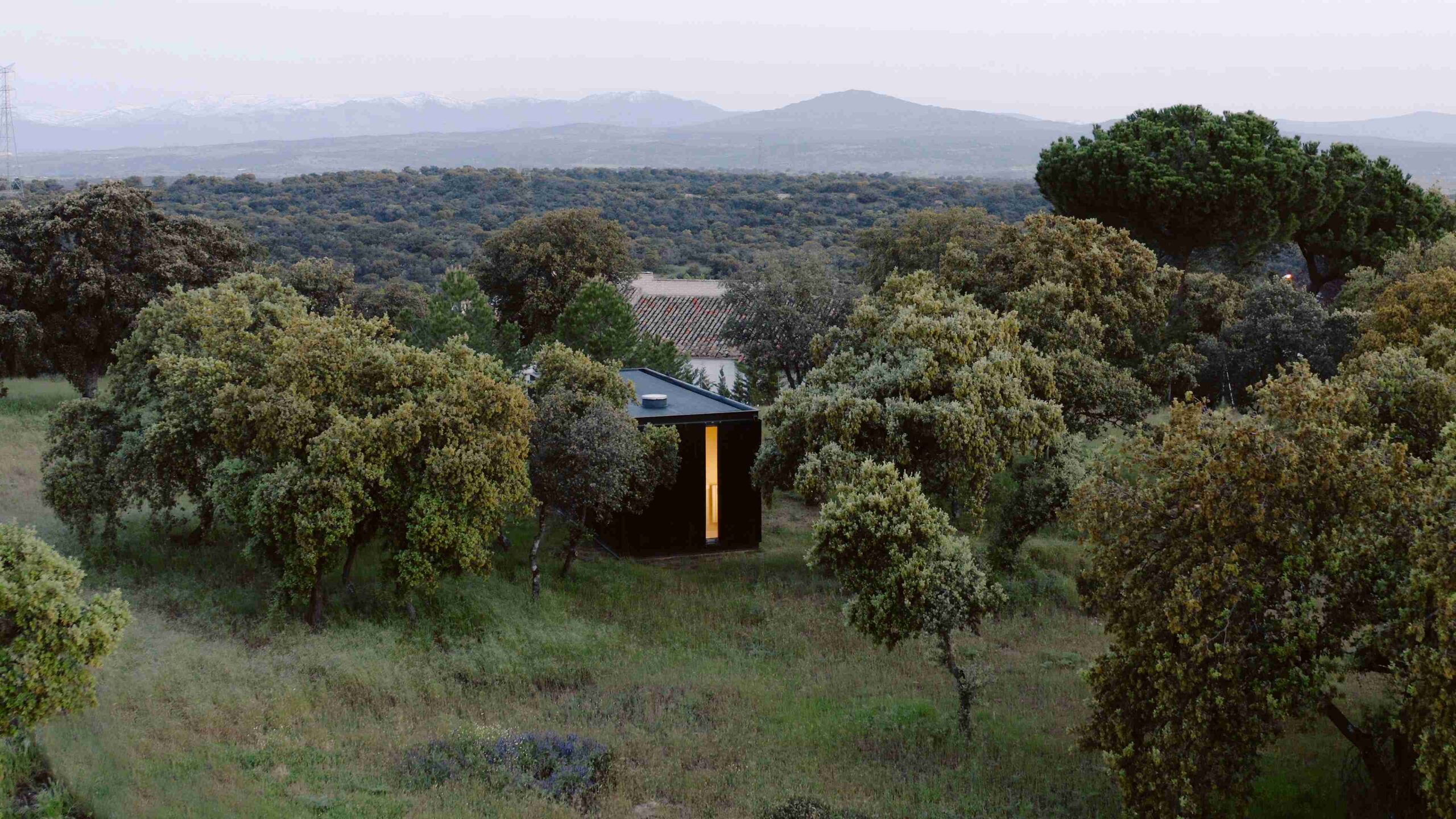 Casa modular Tini XS de fachada negra iluminada por dentro, en un campo de encinas con montañas al fondo